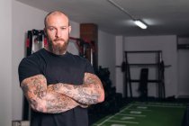 Сильний дорослий спортсмен з бородою та татуюваннями на складених руках дивиться на камеру в гімназії — стокове фото