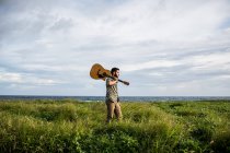 Hombre tranquilo músico en ropa casual de pie con guitarra acústica en el hombro entre la hierba verde en la costa del océano en verano a la luz del día - foto de stock