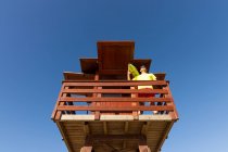 Low angle of brave lifeguard in sunglasses standing on wooden rescue tower and monitoring safety under blue sky — Stock Photo
