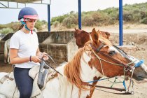 Side view of girl in helmet and casual outfit sitting in saddle on horse with bridle on sandy meadow near grass in nature in daylight — Stock Photo