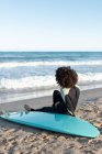 Вид ззаду повне тіло анонімного босоногих жіночого серфера в гідрокостюмі, що сидить на піщаному пляжі біля моря, що махає — стокове фото