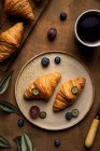 Сверху вкусные свежие круассаны подаются на тарелке с фруктами, помещенными возле чашки чая на деревянном столе в утреннее время в светлом помещении — стоковое фото