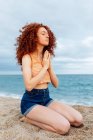 Side view full body of peaceful female with flying curly hair sitting on knees on sandy seashore and making namaste gesture during meditation — Stock Photo