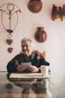 Donna anziana sorridente che indossa vestiti caldi seduta a tavola con tablet e tazza di tè distogliendo lo sguardo — Foto stock