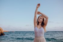 Attractive young female with long hair and closed eyes while standing on seashore in summer on blurred background with outstretched arms — Stock Photo