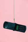 Vieux téléphone portable flip ouvert noir accroché à un crochet métallique avec corde verte sur fond rose dans un studio moderne et lumineux — Photo de stock