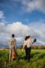 Back view of unrecognizable friends musicians walking with guitars on pathway among green grass on coast near ocean in daytime — Stock Photo
