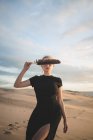 Serious female covering her eyes with black feather wearing dress standing on sandy dune washed by sea at sundown — Stock Photo