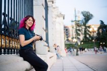 Fotógrafa positiva con cabello rosa y cámara fotográfica en la mano mirando la cámara mientras está sentada cerca de la cerca del edificio envejecido en la plaza - foto de stock