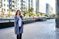 Positive female with long straight fair hair in eyeglasses and formal wear recording voice message while standing on street — Stock Photo