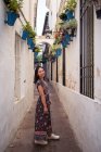 Ganzkörper-Seitenansicht einer lächelnden asiatischen Touristin, die in einem schmalen Gang zwischen Häusern steht, während sie in die Kamera in der Stadt blickt — Stockfoto