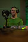 Positif jeune homme barbu millénium en lunettes et écouteurs souriant et parlant en micro tout en enregistrant podcast en studio sombre — Photo de stock