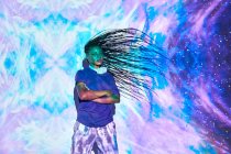 Seitenansicht der ethnischen weiblichen Millennial schüttelt lange geflochtene Haare, während seine Arme in der Nähe der Wand mit leuchtend blauen und lila Beleuchtung gekreuzt sind — Stockfoto