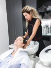 Professionnel sérieux femme coiffeur laver les cheveux du client dans l'évier avec robinet dans le salon de coiffure près du comptoir avec des bouteilles et des étagères avec divers outils — Photo de stock