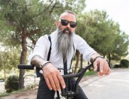 Confiado hipster masculino con tatuajes en camisa blanca y gafas de sol sentado en bicicleta en el parque con árboles verdes en la ciudad - foto de stock