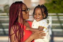 Allegro madre afro-americana con trecce rosse in piedi con figlia positiva sulle mani sulla strada alla luce del sole — Foto stock