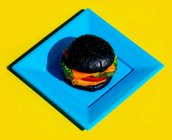 Зверху свіжий бургер з чорною булочкою та овочами, поданими на квадратній синій тарілці на жовтому фоні — стокове фото