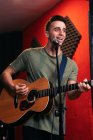 Positiver junger männlicher Gitarrist spielt Akustikgitarre und singt im Lichtclub am Mikrofon — Stockfoto