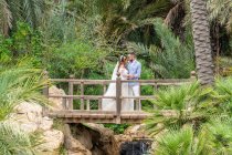 Молодая пара в свадебных нарядах, стоящая на деревянном пешеходном мосту с перилами и держась за руки, обнимаясь и глядя друг на друга над водопадом со скалами возле зеленых деревьев в парке — стоковое фото