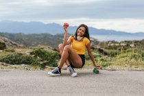 Full body of positive ethnic female with wireless headphones on neck and apple in hand while sitting on longboard on asphalt road — Stock Photo
