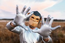 Anonymous person wearing geometric monkey mask while demonstrating hands with bent thumbs at camera against blurred background of countryside — Stock Photo