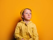 Unbeweglicher kleiner Junge in trendigem Regenmantel und Mütze, der vor gelbem Hintergrund im Atelier wegschaut — Stockfoto