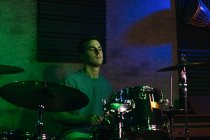 Concentrated young male musician playing drums in club with green and blue neon illumination — Stock Photo