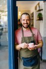 Barista masculino barbudo positivo en delantal con taza de café caliente en las manos de pie en la puerta de la cafetería moderna - foto de stock