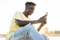 Lächelnder afroamerikanischer Mann in lässiger Kleidung sitzt an felsiger Küste, während er im Sommerabend sein Smartphone benutzt — Stockfoto