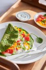 Overhead of spinach creppe with tomato and pepper slices served on white dishware on tray near salad leaves and sour cream — Stock Photo