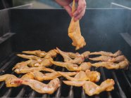 Crop chef anônimo colocando asa de frango cru na grelha de metal quente com fumaça enquanto cozinha no campo durante o piquenique — Fotografia de Stock