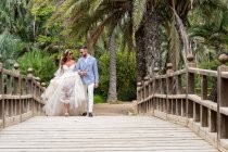 Brautpaar in Hochzeitskostümen läuft auf Holzsteg mit Geländer und hält Händchen und schaut sich an grünen Palmen und Pflanzen im Garten im Sommertag an — Stockfoto