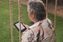Visão traseira da senhora idosa vestindo blusa sentada no parque e lendo livro eletrônico — Fotografia de Stock