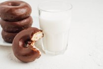 Mucchio di ciambelle dolci e gustose con glassa di cioccolato posta sul piatto sul tavolo bianco con bicchiere di latte nella stanza luminosa — Foto stock
