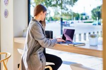 Seitenansicht einer konzentrierten Geschäftsfrau, die auf einem Stuhl sitzt und auf einem Netbook tippt, während sie im Café an einem Remote-Projekt arbeitet — Stockfoto