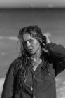 Black and white of female in wet shirt and with wet hair standing looking at camera on beach near sea while enjoying summer day — Stock Photo