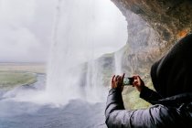 Side view of unrecognizable traveler in warm outerwear and hoodie taking photo of picturesque Seljalandsfoss waterfall on smartphone during trip in Iceland — Stock Photo