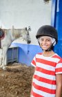 Lächelndes Kind in Jockeymütze und Freizeitkleidung, das bei Tageslicht in der Nähe eines weißen Pferdes im Stall neben einer Hauswand steht und in die Kamera blickt — Stockfoto