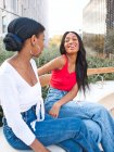 Ernsthafte afroamerikanische Freundinnen reden und schauen einander an, während sie auf einer Bank in der Nähe grüner Pflanzen auf einer Straße mit Gebäuden sitzen — Stockfoto