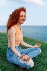 Side view of carefree female with long ginger hair sitting looking at camera in Padmasana pose with zen gesture on grassy hill on seashore — Stock Photo