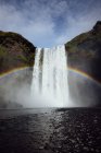 Impresionante paisaje de arco iris brillando sobre la rápida y poderosa cascada de Skogafoss que fluye a través de un acantilado rocoso en Islandia - foto de stock