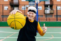 Femme mature positive en vêtements de sport et bandeau regardant la caméra tout en se tenant debout avec la balle dans le bras tendu pendant le match de basket-ball — Photo de stock