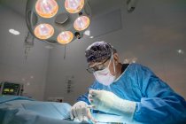 Профессиональный старший хирург в маске и форме делает операцию под лампой в операционной — стоковое фото