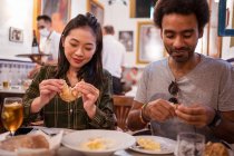 Feliz joven pareja multiétnica en ropa casual sonriendo mientras come deliciosos alimentos durante la fecha en el restaurante moderno - foto de stock