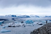 Vista lateral de excursionista anónimo distante en ropa interior caliente y sombrero de pie en la colina rocosa y admirar impresionantes paisajes de Jokulsarlon gran lago glaciar durante el viaje en Islandia - foto de stock
