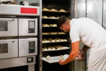 Young stylish tattooed male baker in casual clothes and eyeglasses putting loaves of raw dough on metal rack while baking bread in kitchen — Stock Photo
