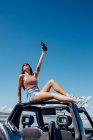 Corps complet de jeune femme heureuse en tenue d'été et lunettes de soleil levant la main de la bière tout en étant assis sur le toit de la voiture safari sur le rivage de la rivière — Photo de stock