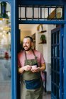 Barista masculino barbudo positivo em avental com xícara de café quente em mãos em pé na porta do café moderno — Fotografia de Stock