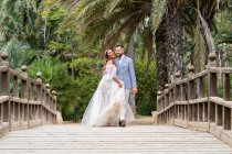 Ehepaar in Hochzeitskostümen steht auf hölzerner Fußgängerbrücke mit Geländer, während es sich umarmt und bei grünen Palmen und Pflanzen im Garten an Sommertagen wegsieht — Stockfoto