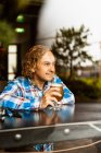 Задумчивый мужчина с вьющимися волосами в повседневной одежде сидит у деревянного прилавка возле окна в баре и пьет пиво днем — стоковое фото